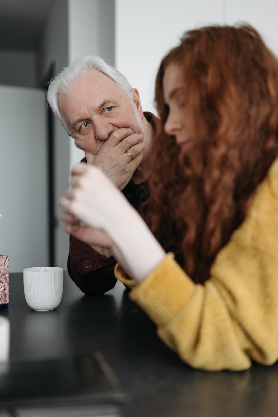 An elderly man and a young woman share a thoughtful conversation at a kitchen countertop.