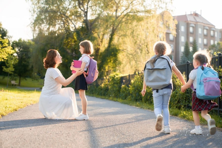 mother wearing long white skirt walking her three daughters to school with backpacks on