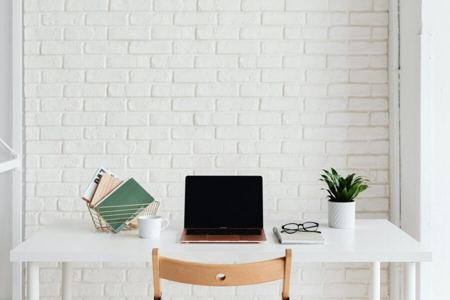 Clean and organized white workspace with laptop, plant, and books against a brick wall.