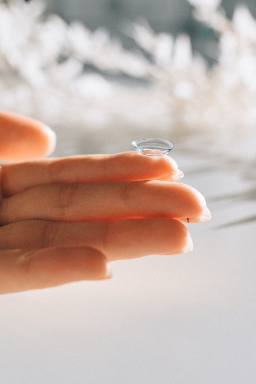Close-up photograph of a hand holding a clear contact lens with a blurred background.