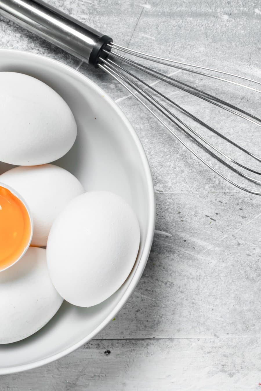 eggs in a white bowl with a metal whisk laying nearby on a grey tabletop