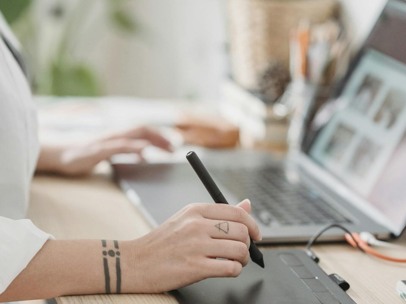 Crop anonymous female browsing netbook and working on graphic tablet with stylus while sitting at desk