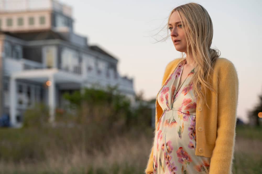 This image shows Dakota Fanning portraying Abby Winbury in The Perfect Couple. Standing in front of a grand coastal home, Abby's serene and contemplative expression, combined with her flowing floral dress, highlights her character's emotional depth and the central role she plays in the unfolding drama.
