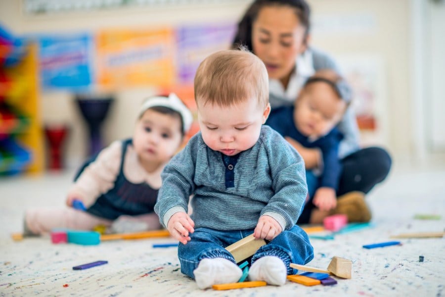 A baby boy is sitting in front of other babies and teacher at a daycare center. He is playing with colorful toy blocks.