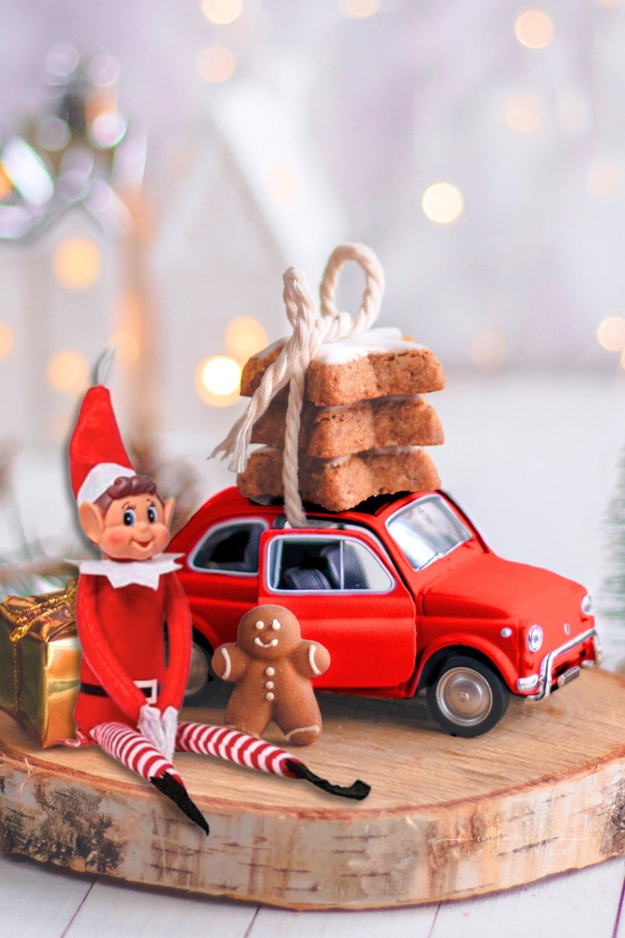Christmas Elf sitting on wood cirle next to red toy car with gingerbread cookies on top