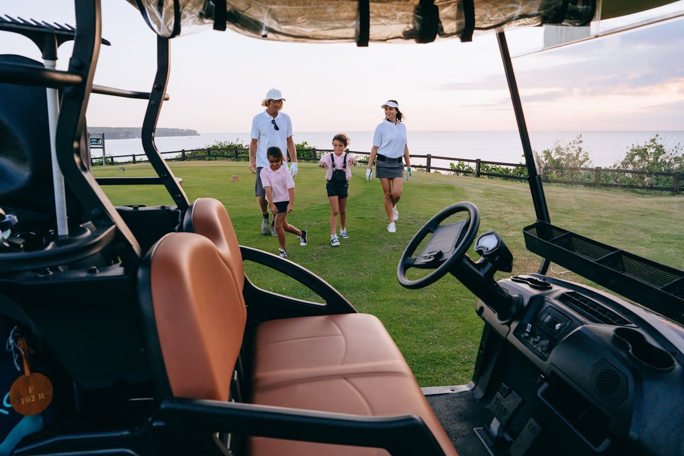 Family enjoying a sunny day walking on a scenic golf course near a golf cart.