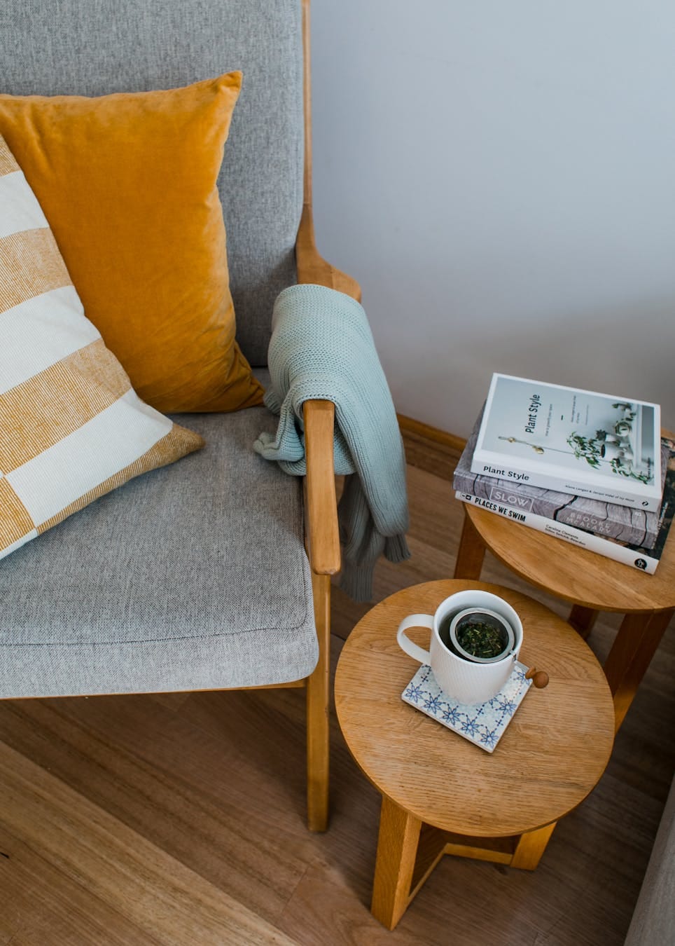 From above of comfortable armchair with colorful pillows placed near wooden side tables with books and cup of tea in living room