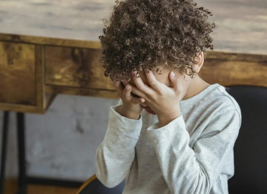 From above of crying African American child with curly hair wearing casual clothes sitting in chair near table and covering e