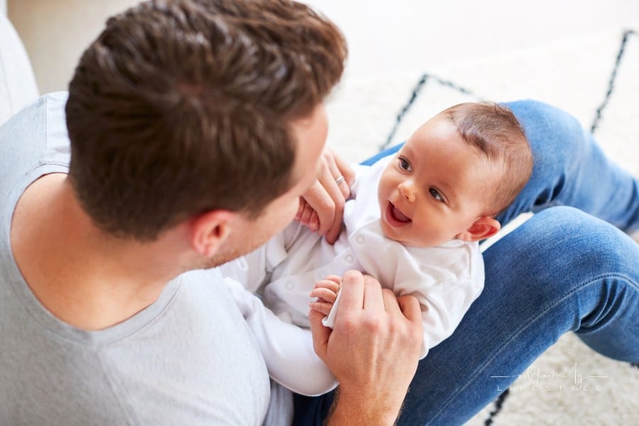 Baby Daughter Lying On Fathers Lap As He Plays Game With Her At Home