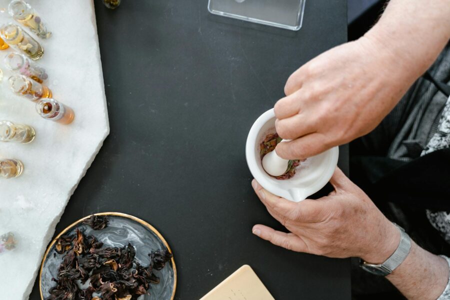 Hands preparing an herbal mixture using a mortar and pestle with dried flowers and oils.