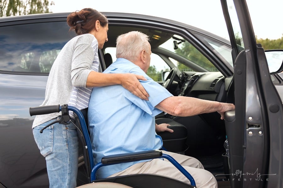 adult daughter helping aging father out of wheelchair and into vehicle