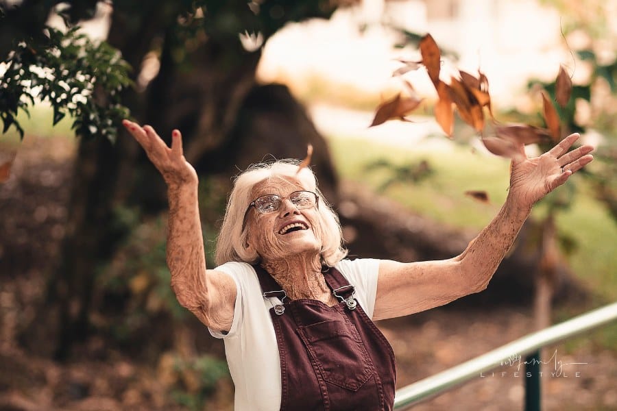 Elderly Woman Wearing Brown Overalls Near Fall Tree throing leaves in the air