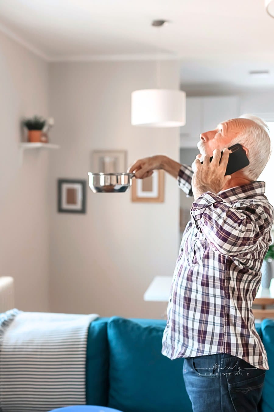 Angry Bearded Senior Man Talking on Smartphone Near Plastic Wash Bowl at Home in the Living Room Because of Roof Leaking