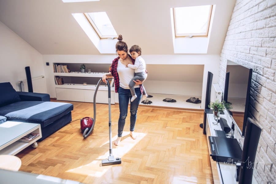 Young woman using vacuum cleaner to clean house and carryng her daughter