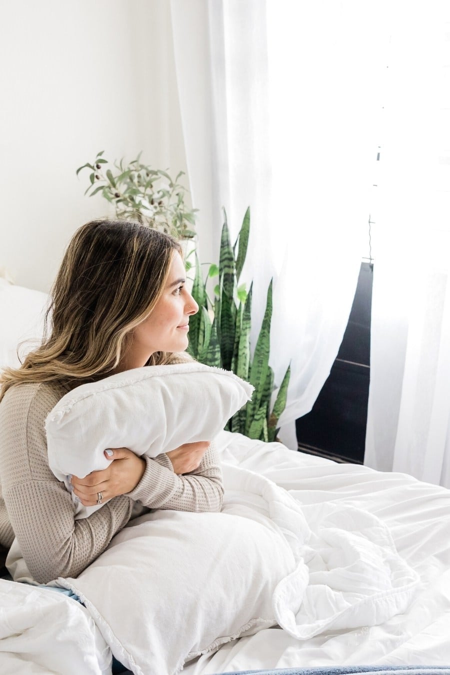 woman holding pillow while sitting on bed and looking out window