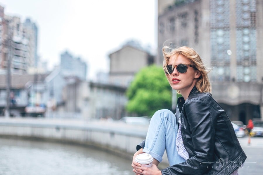 young woman in leather jacket and light colored jeans sitting by pond drinking coffee