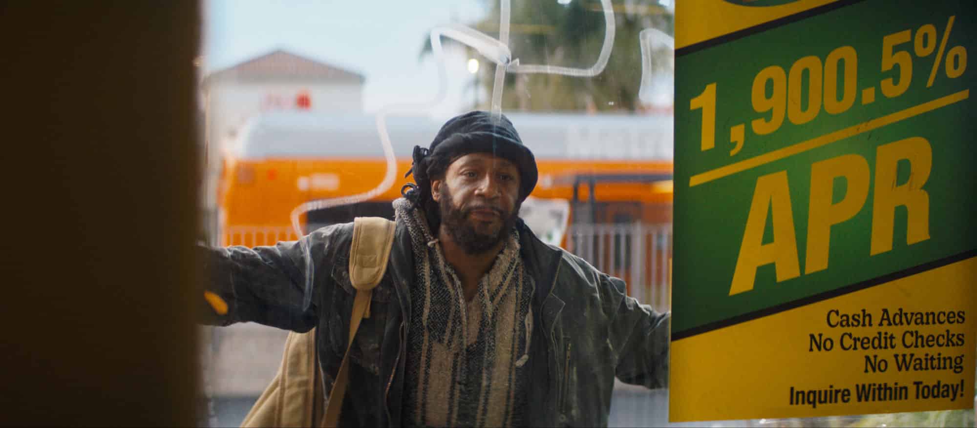 Katt Williams stares through a payday loan store window, looking worn and serious. Description: Comedian and actor Katt Williams in One of Them Days, seemingly playing a streetwise character with a keen eye on survival in an urban setting.