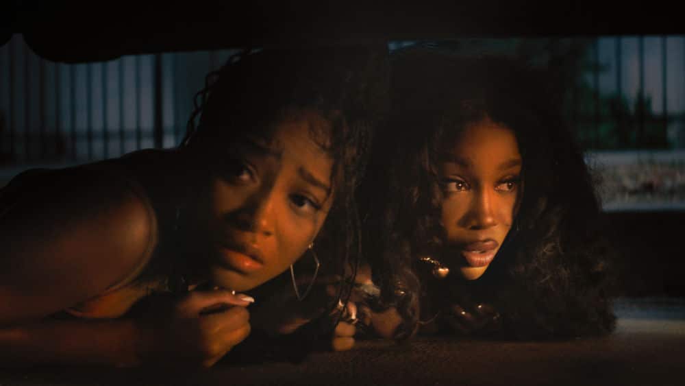 Keke Palmer and SZA crouch under a car, looking tense and concerned.