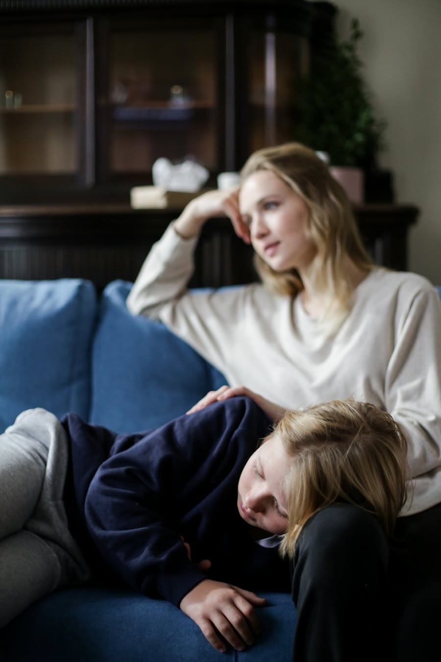Mother with a thoughtful expression comforts her resting child on a cozy couch.