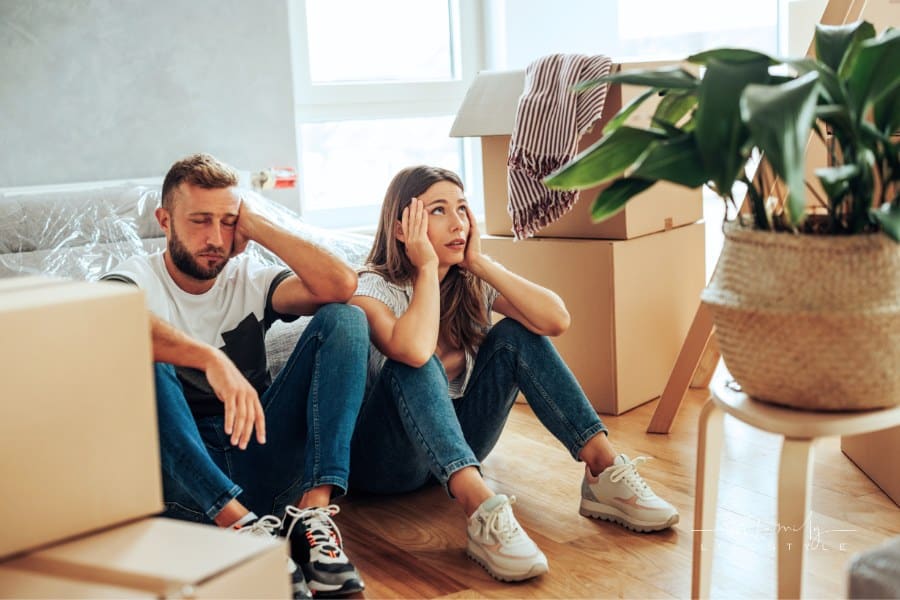 A Caucasian couple is sitting on the floor of their apartment. All around them are packed boxes and they are very stressed be