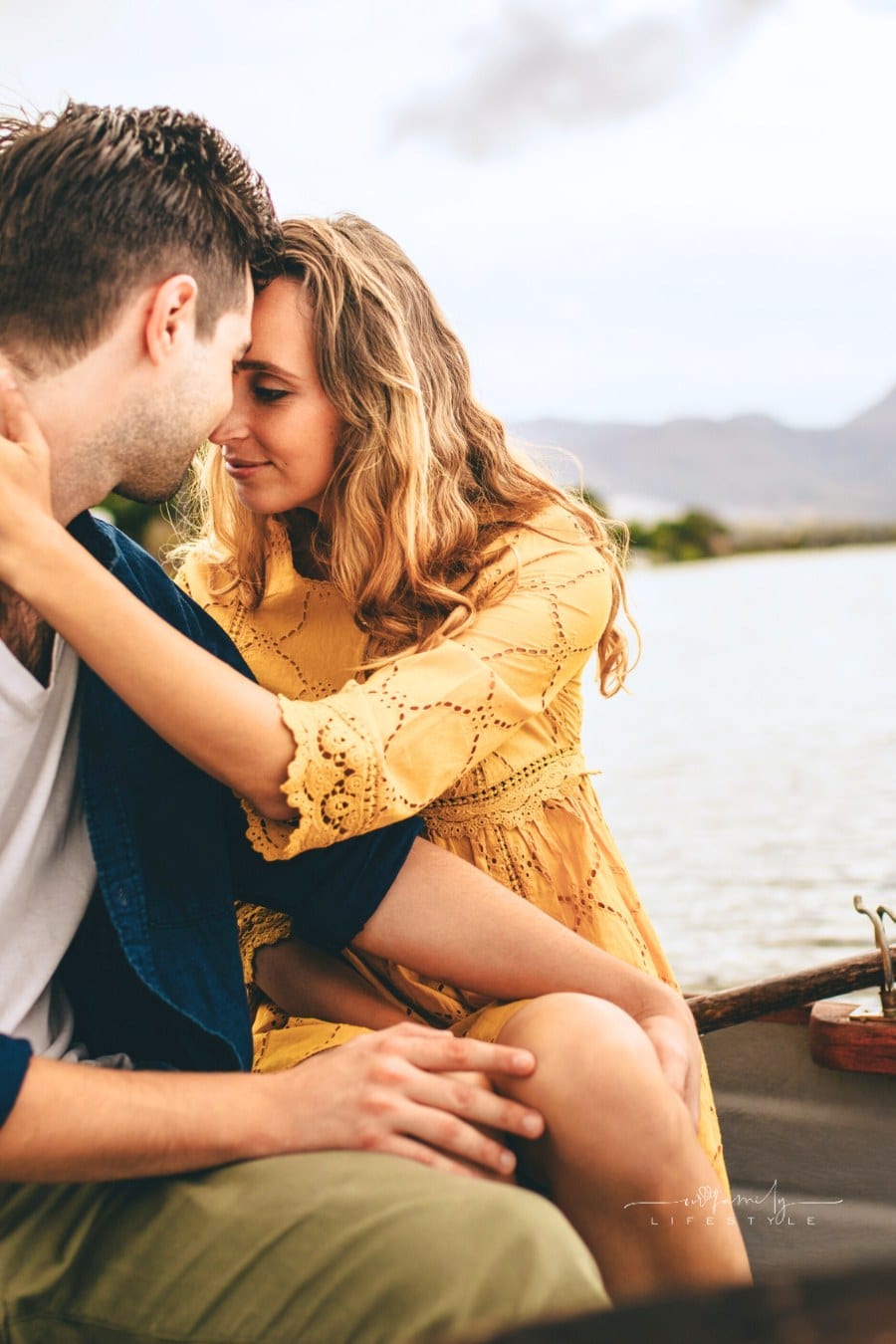 Young couple sitting together in a boat touching their heads. Couple in love on a boat date in a lake with hills and houses in the background.
