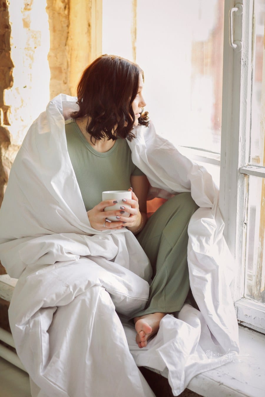 Relaxed positive young brunette woman in homewear sitting on top of unmade bed with cup of tea in her hands, slowly drinking aromatic hot beverage while enjoying spending free time alone at home