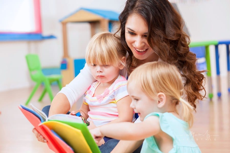 daycare teacher sitting on the floor and reading a colorful book to two preschool girls. The little girls are interested in t