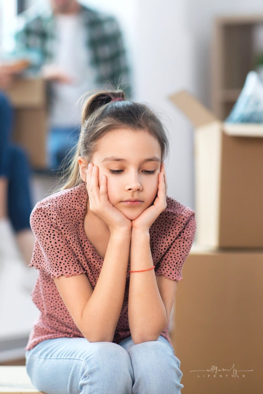 sad girl sitting on moving box sad about moving to new home while parents talk behind her