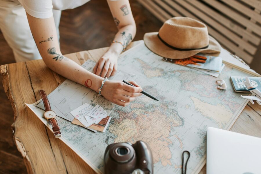 Tattooed woman planning a trip with a world map, passports, and travel essentials on a wooden table.