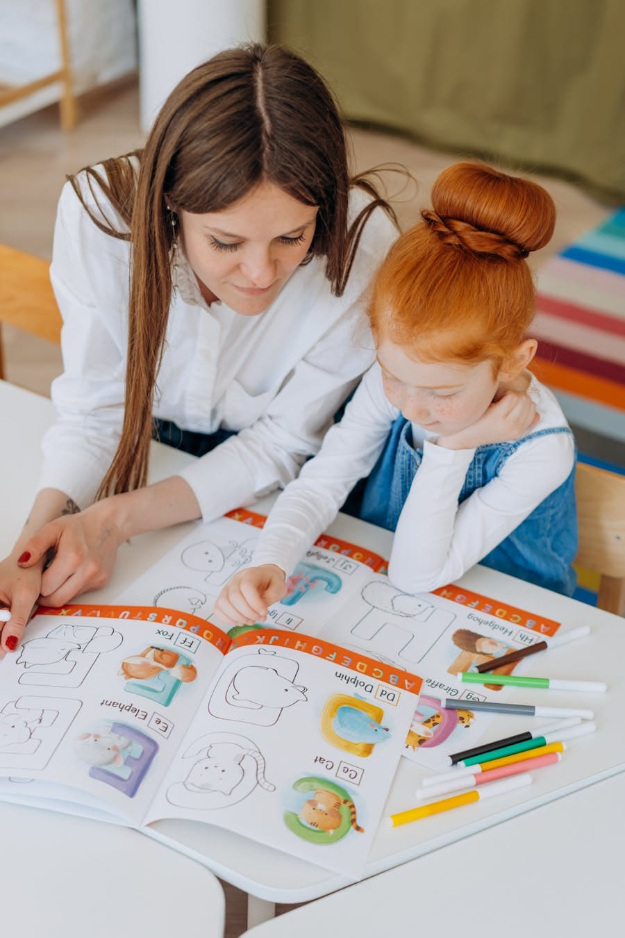 Teacher helping a young girl with her alphabets in a colorful classroom setting.