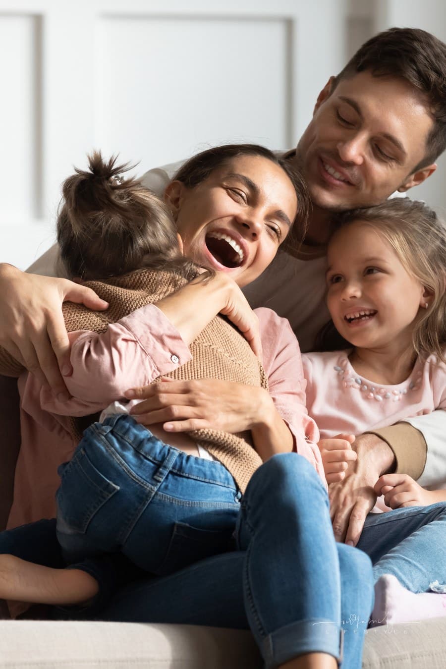Cheerful young father and small daughter watching preschool daughter cuddling with affectionate mother. Happy family having fun, sitting together on comfortable couch in living room.