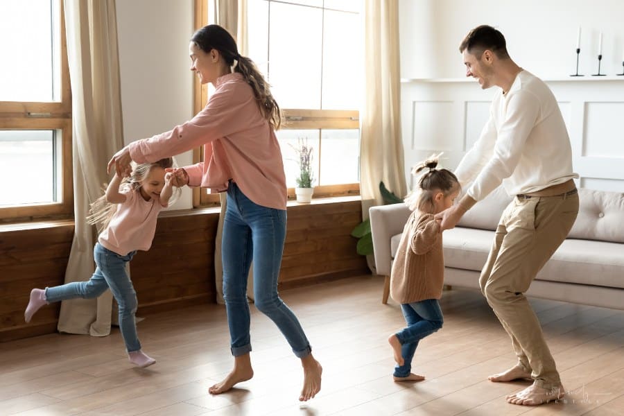 Happy active family young mom dad and cute little children daughters holding hands dancing together in living room interior, carefree funny small kids having fun jumping laughing with parents at home