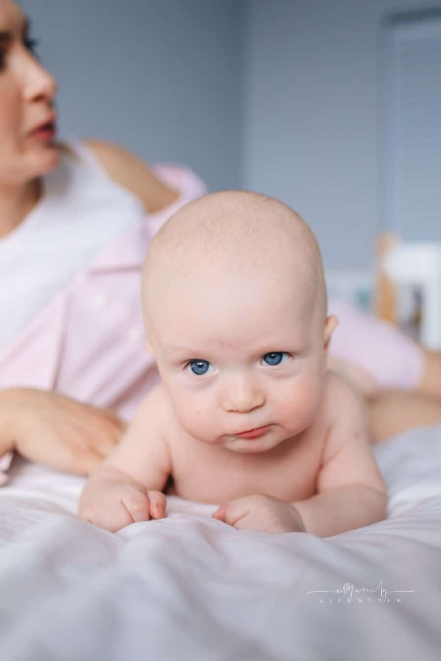 Blue-eyed Baby laying on stomach on bed with mom behind her