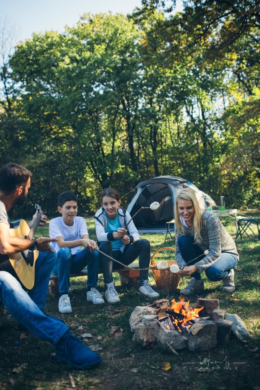 father playing guitar while mom and two kids roast marshmallows over fire for s'mores