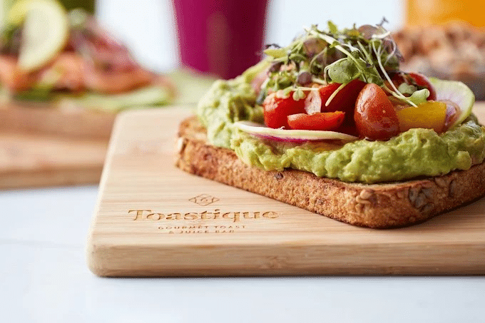Close-up of gourmet avocado toast topped with cherry tomatoes, onions, and microgreens, served on whole-grain bread at Toasti