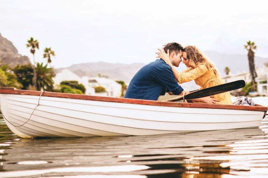 Young couple sitting together in a boat touching their heads. Couple in love on a boat date in a lake with hills and houses i