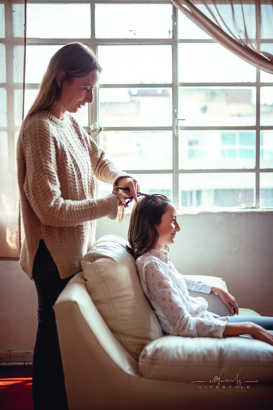 side view of mother braiding daughter's hair in front of a large window