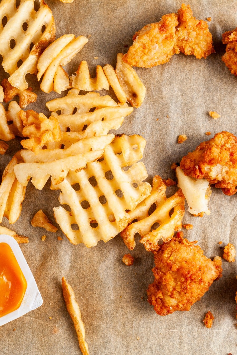 flat lay image of a messy food with waffle fries and chicken nuggets on baking paper together with ketchup and other sauces and dips. Some are eaten others dipped. A messy junk food scene.