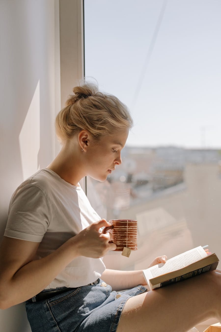 Woman enjoying a book and tea by a window, capturing a peaceful indoor moment in natural light.