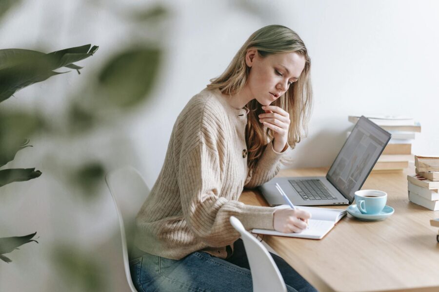 Woman sitting at a desk writing notes, using laptop, with coffee cup.