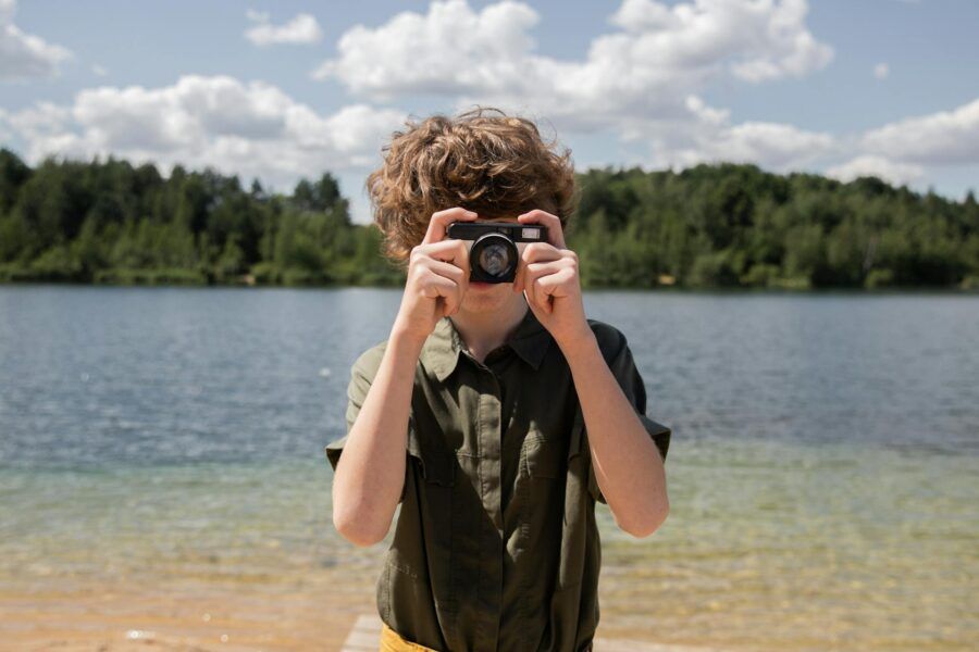 A boy with curly hair taking pictures by a lake on a bright sunny day.