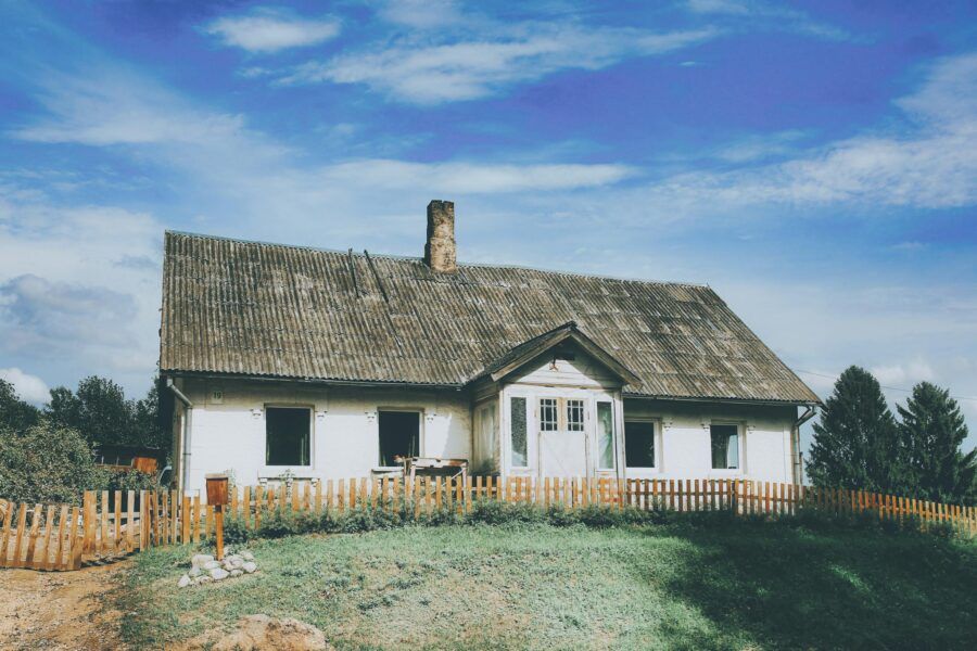 A rustic abandoned house with a weathered facade under a bright blue sky, surrounded by greenery.