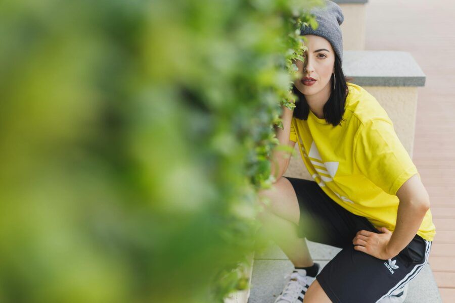 A woman in a yellow Adidas shirt squats near a hedge, showcasing streetwear style.