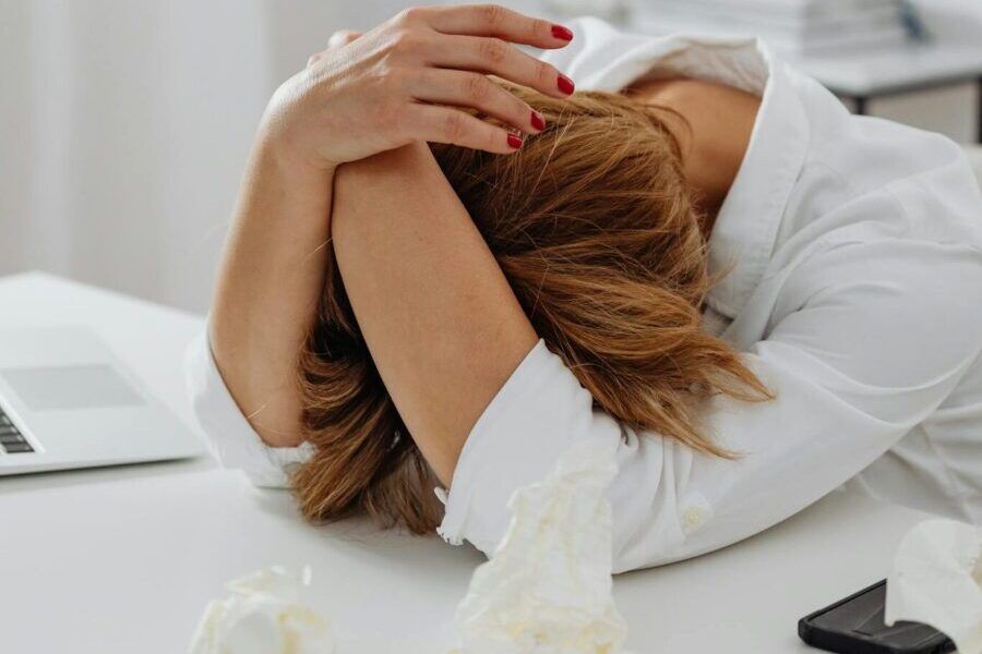 A woman in distress sits at a desk with head in hands, surrounded by tissue papers.