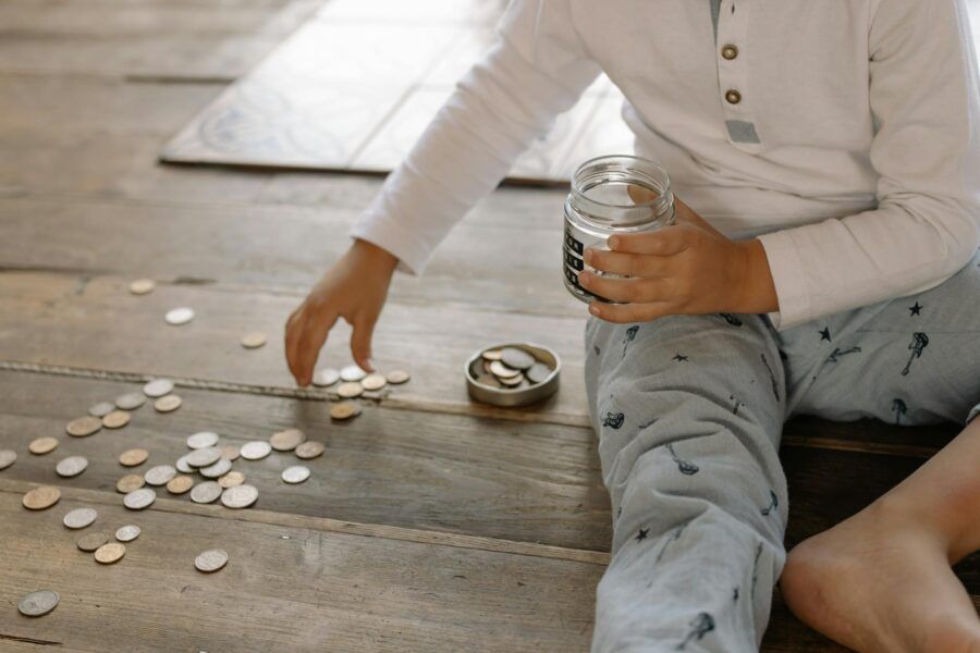 A young child collects coins into a jar on a wooden floor, symbolizing savings.
