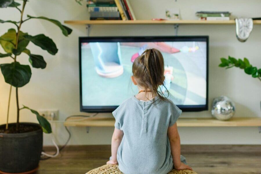Back view of unrecognizable little kid watching television in living room with potted plants in house