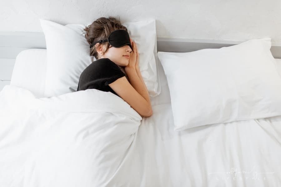 woman lying with a sleeping mask in bed under white comforter blanket