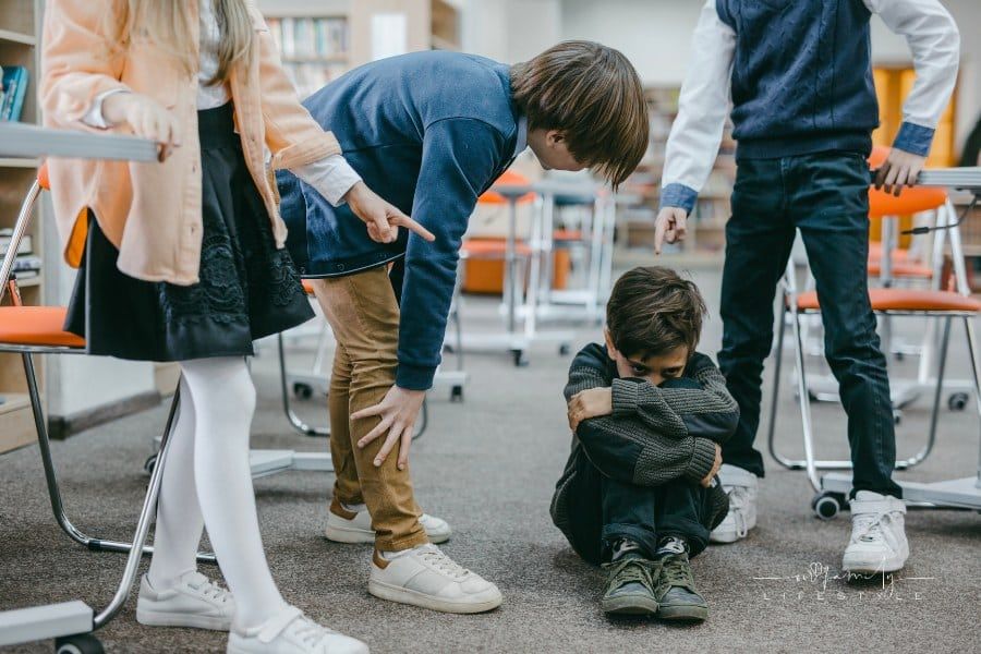 A Boy in Gray Sweater Sitting on the Floor while other kids point and bully him