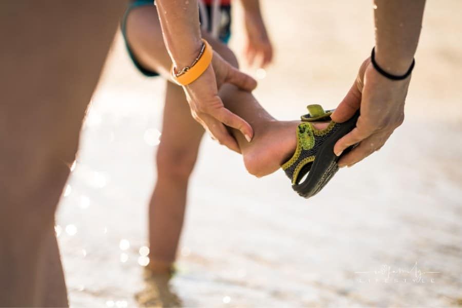 Close up of mother assisting her child with putting on aqua shoes at sea.
