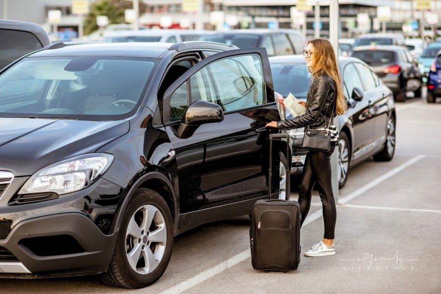 young and happy woman traveling with suitcase near the rental car outdoors on the parking